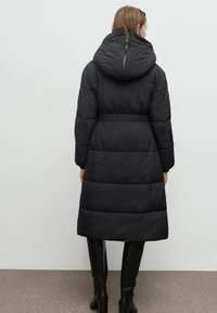 Mujer con un largo abrigo negro acolchado con capucha, de pie de espaldas, combinado con botas negras hasta la rodilla.