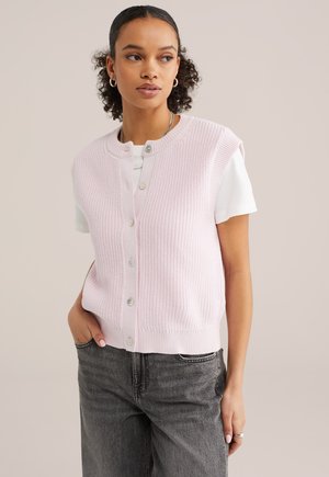 Giovane donna con capelli ricci indossa un gilet lavorato a maglia rosa chiaro con bottoni sopra una t-shirt bianca e jeans grigio scuro, posando con una mano in tasca.