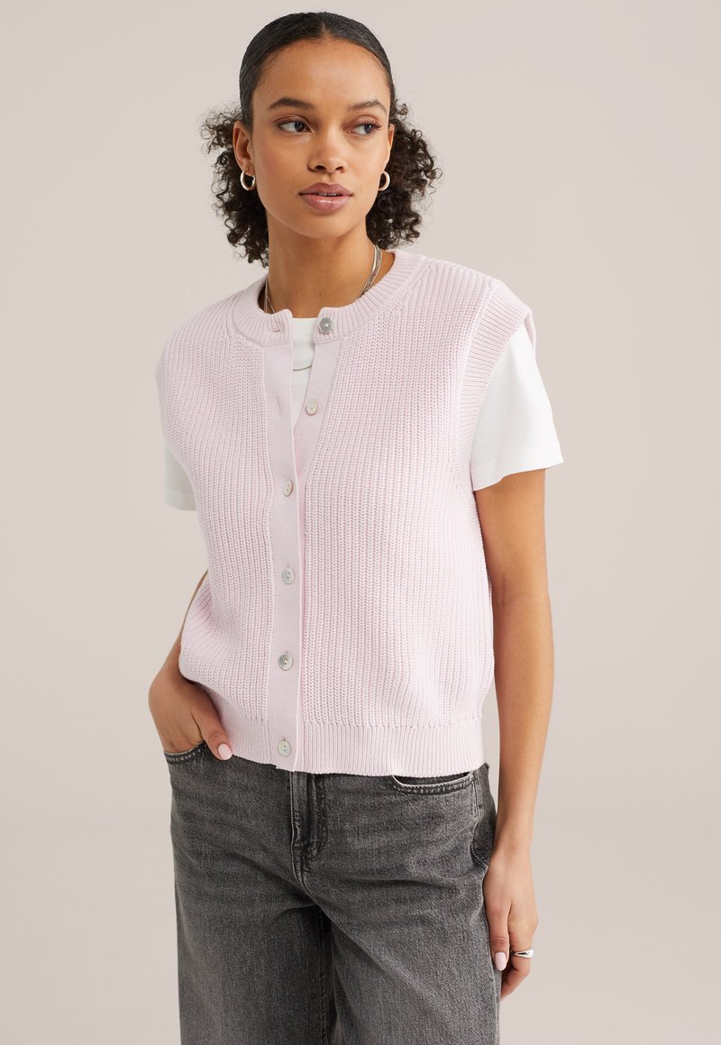 Mladá žena s kučeravými vlasmi má na sebe svetloružovú pletenú vestu na gombíky cez biele tričko a tmavosivé džínsy, pózuje s jednou rukou v vrecku.