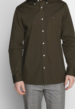 Mies, jolla on päällään tummanoliivinvärinen pitkähihainen nappipaita, jossa pieni logo rinnassa, ja harmaat ruudulliset housut yksiväristä taustaa vasten.