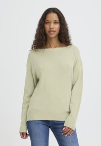 Pull vert clair en maille douce avec un large décolleté, des poignets côtelés et des manches longues, porté avec un jean bleu.