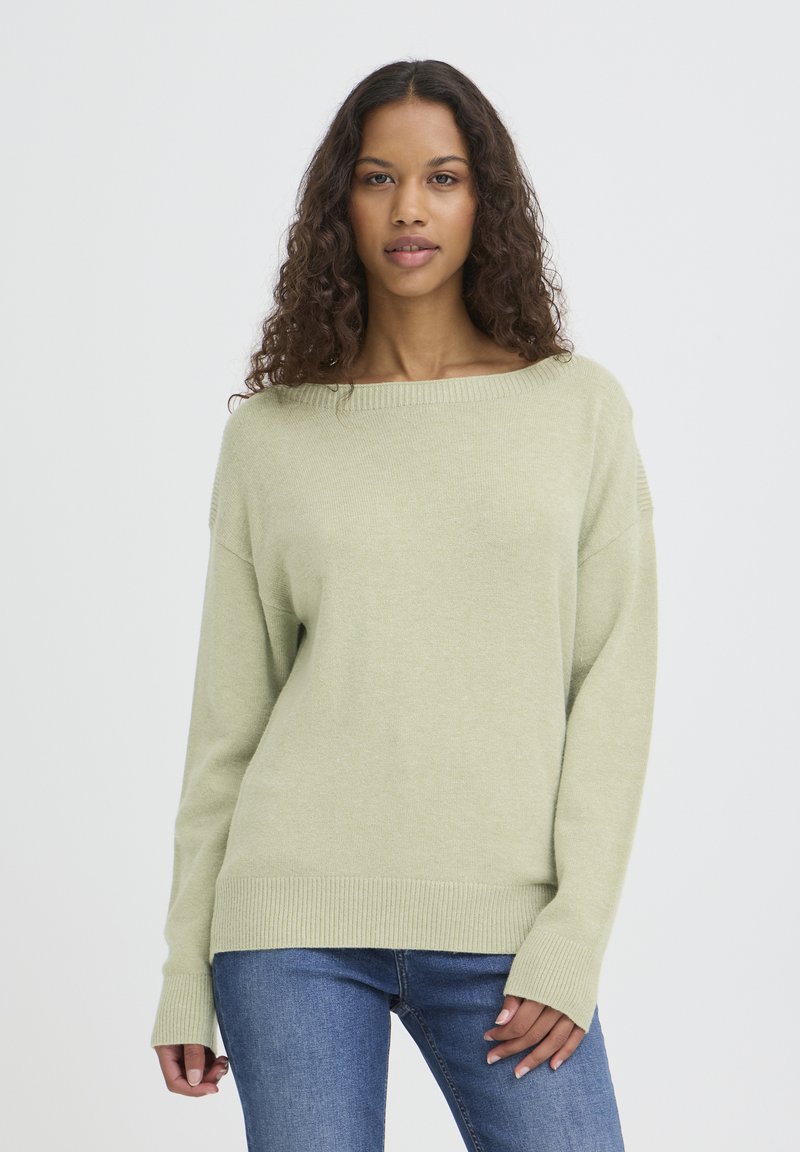Pull vert clair en maille douce avec un large décolleté, des poignets côtelés et des manches longues, porté avec un jean bleu.