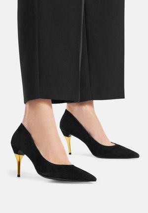 Scarpe nere in suede con tacco alto, punta appuntita e tacco di 7 cm con dettagli dorati, abbinate a pantaloni neri su misura.