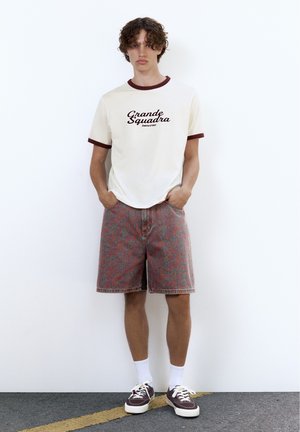 Tânăr stă sprijinit de un perete alb, purtând un tricou crem cu text, pantaloni scurți cu model până la genunchi, șosete albe și adidași maro.