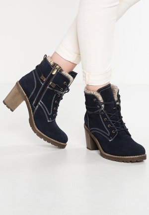 Dunkelblaue Wildleder-Stiefeletten mit klobigen Absätzen, Fellfutter, seitlichen Reißverschlüssen und Schnürung an der Vorderseite, getragen zu hellen Hosen.