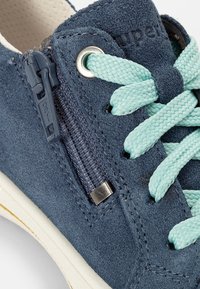 Blaue Wildleder-Sneaker mit einem seitlichen Reißverschluss, mintgrünen Schnürsenkeln und weißer Sohle. Texturierte Oberfläche mit sichtbaren Nähten und Metallakzenten.