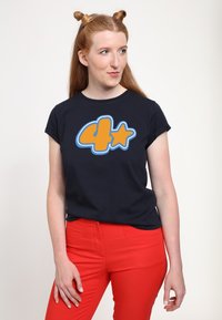 Marineblaues T-Shirt mit einer großen orangefarbenen "4" und einem Stern-Graphic, aus Baumwolle, mit kurzen Ärmeln und lockerer Passform. Kombiniert mit roten Hosen.