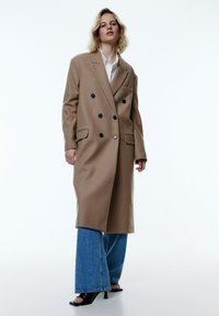 Dubbelzijdig beige jasje met zwarte knopen, zijzakken en een relaxte pasvorm, gedragen over lichtblauwe flare jeans en zwarte open hakken.