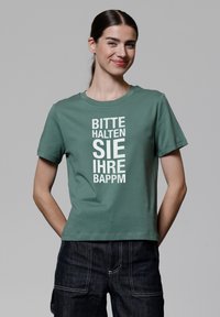 Grünes Baumwoll-T-Shirt mit kurzen Ärmeln und Rundhalsausschnitt. Auf der Vorderseite befindet sich fett gedruckter weißer Text, der vertikal angeordnet ist. Kombiniert mit dunklen Jeans.
