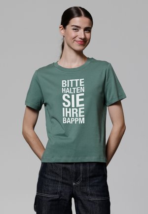 Grünes Baumwoll-T-Shirt mit kurzen Ärmeln und Rundhalsausschnitt. Auf der Vorderseite befindet sich fett gedruckter weißer Text, der vertikal angeordnet ist. Kombiniert mit dunklen Jeans.