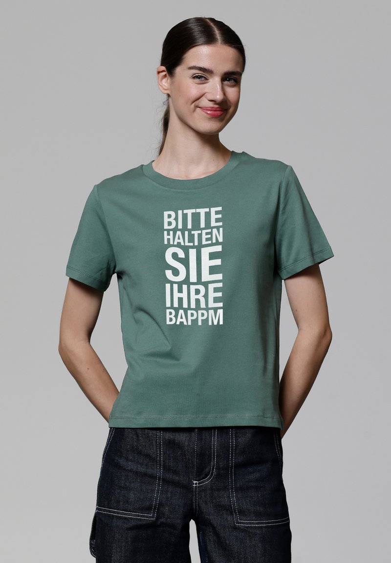 Grünes Baumwoll-T-Shirt mit kurzen Ärmeln und Rundhalsausschnitt. Auf der Vorderseite befindet sich fett gedruckter weißer Text, der vertikal angeordnet ist. Kombiniert mit dunklen Jeans.