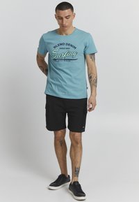 Svijetloplavi pamučni majica s natpisom "Blend Denim Surfing". Crne cargo kratke hlače. Model nosi crne tenisice, prikazujući ležeran izgled.