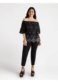 Top negro sin hombros con patrones bordados en blanco, mangas acampanadas y dobladillo con forma de pico, combinado con pantalones negros ajustados y zapatos de plataforma.