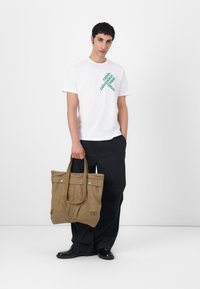Beige lærred tote taske med dobbelte hanke, frontlommer og metal snaplukning. Tasken har et subtilt brandlogo på siden.