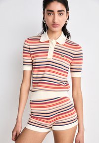 Ensemble polo tricoté rayé et shorts en crème, rouge, bleu et violet. Comprend un col, des boutons et un logo sur le côté gauche de la poitrine.