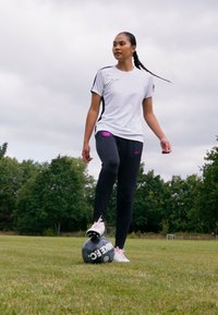 Atletka vo bielej športovej košeli a čiernych legínach, stojaca na čiernej futbalovej lopte, má na sebe červené a biele športové topánky, trávnaté pole.