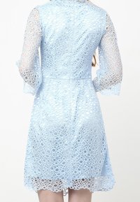 Ljusblå klänning i intrikat spets med cirkulära utskärningar. Har långa, utsvängda ärmar och vågiga kanter vid nederkanten.