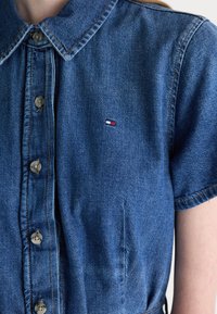 Denim shirt with a light blue color, short sleeves, button front, and a small embroidered logo featuring red and white accents on the left chest.