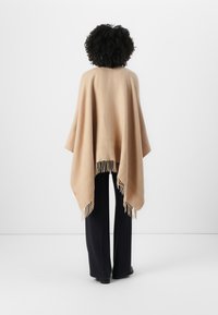 Beige poncho med frynser, laget av mykt stoff. Løs design med trekantet form, brukt over marineblå bukser.