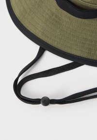Olive green bucket hat with a textured material, wide brim, and black accents. Includes adjustable black cords with a toggle for a secure fit.