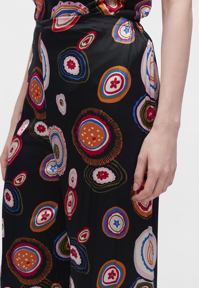 Gros plan d'une personne portant un pantalon noir avec un motif floral circulaire coloré et sa main droite détendue le long du corps.