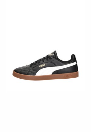 Zapatilla negra de Puma con franja blanca, logo de Puma beige, cordones negros y suela marrón, vista desde el lado exterior.
