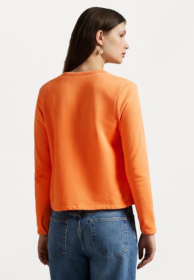 Orange long-sleeve shirt with ribbed texture, crew neck, and cropped hem. Paired with high-waisted blue denim jeans.