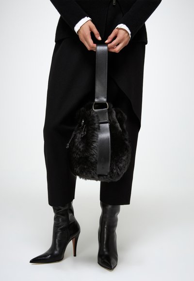 Borsa a secchiello in pelliccia nera con una tracolla in pelle, dotata di un cinturino regolabile e dettaglio del brand. Indossata con stivali neri con tacco alto e pantaloni.