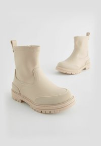 Bottines beiges imperméables avec une texture lisse, bout rond, semelle en caoutchouc et languette à l'arrière. Ils présentent des détails à texture fine.