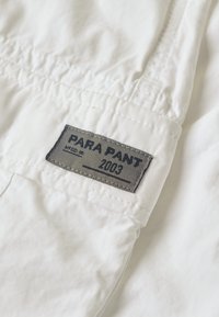 Białe spodnie materiałowe z szarym metkiem z napisem "PARA PANT 2003", które prezentują gładką teksturę i prosty design.