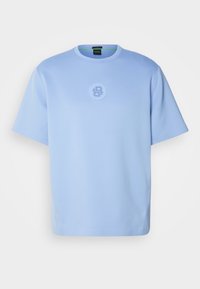 T-shirt bleu clair à manches courtes en tissu lisse. Présente un logo circulaire au centre et un col rond.