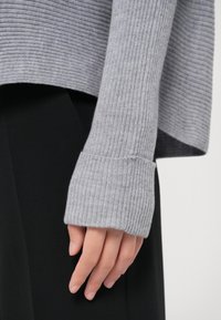 Gros plan d'une main partiellement couverte par une manche de pull en maille côtelée grise avec un large poignet, devant un pantalon noir.