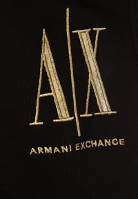Černá látka s výrazným zlatě vyšitým logem obsahujícím text "A/X" a "ARMANI EXCHANGE", představující elegantní a moderní design.