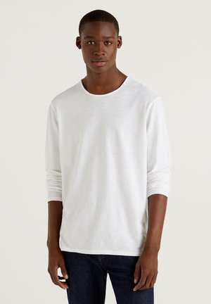 Chemise blanche à manches longues avec une texture lisse, un col rond et des poignets retroussés, associée à un jean bleu foncé.