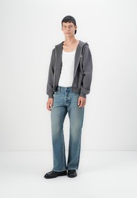 Gray zip-up hoodie, white tank top, light blue jeans with a straight cut, and black shoes. The model wears a dark cap.
