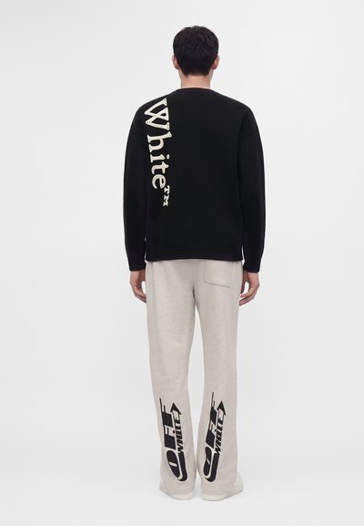 Homme portant un pull noir à manches longues avec le texte blanc "White" et un pantalon beige avec des logos noirs "Off-White" à l’arrière des jambes.