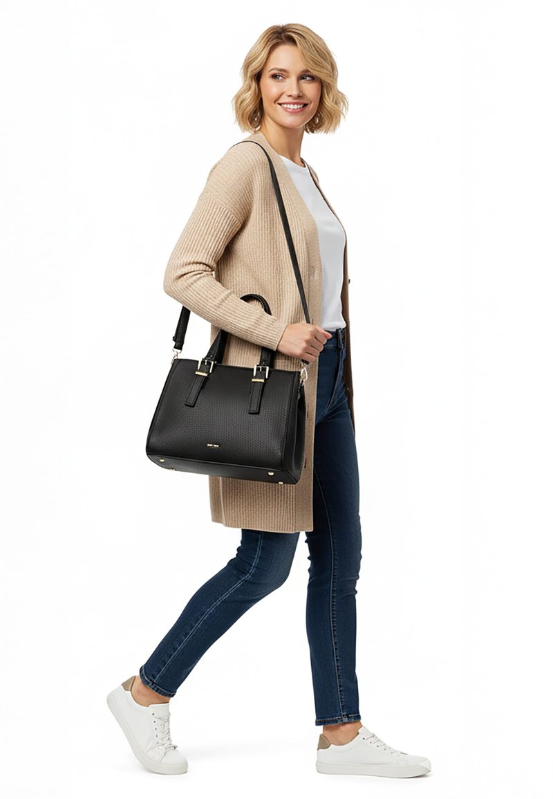 Kobieta idąca, ubrana w beżowy kardigan, białą koszulę, niebieskie dżinsy, białe trampki, niosąca czarną, teksturowaną torebkę z paskami przewieszoną przez ramię.