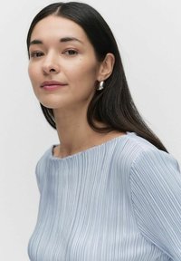 Lyseblå, ribbet stof top med en bred halsudskæring. Glat, lang mørk hår og store sølvøreringe er synlige. Hudtonen fremstår naturlig.