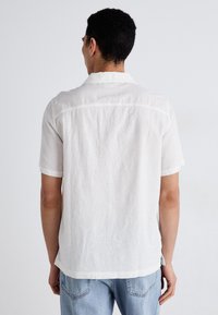 Chemise blanche à manches courtes en tissu léger, avec une coupe décontractée, un col simple et des détails cousus le long du dos.