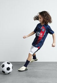 Niño vistiendo una camiseta de fútbol de París en azul marino y rojo con el logotipo de "Catar Airways", pantalones cortos blancos y calcetines negros, pateando un balón de fútbol en blanco y negro.
