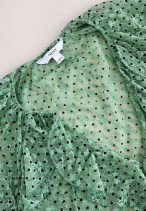 Blusa in pizzo verde con pois neri, tessuto morbido e fluente con vestibilità ampia e etichetta sul collo con la scritta "NEXT".