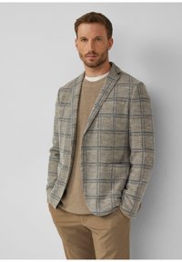 Blazer mit grau-blauem Karomuster, aus einem strukturierten Stoff gefertigt, kombiniert mit einem beigen Pullover und braunen Hosen. Glatte Oberfläche, strukturierter Schnitt.
