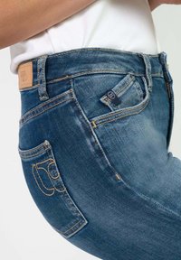 Donkerblauwe denim jeans met een vijfzakkenontwerp, zichtbare stiksels en een leren label aan de tailleband. Zachte textuur met een lichte vervaging.