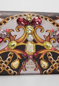 Portafoglio in pelle nera con gemme vivaci in rosso, giallo e viola. Sfondo con un intricato motivo a catena dorata e logo "GUESS EST 1981".