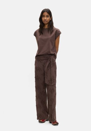 Kobieta o długich ciemnych włosach nosi brązowy bezrękawnik i dopasowane szerokie spodnie o kwiatowym wzorze z przewiązanym paskiem, stojąc na jednolitym tle.