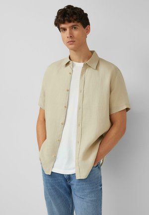Jonge man met krullend donker haar, draagt een beige kortemouwen blouse over een wit T-shirt en blauwe jeans, handen in de zakken.