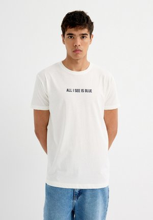 Camiseta de algodón en color blanco roto con mangas cortas, escote redondo y el texto en marino "TODO LO QUE VEO ES AZUL" en el frente. Combinada con pantalones vaqueros azules.