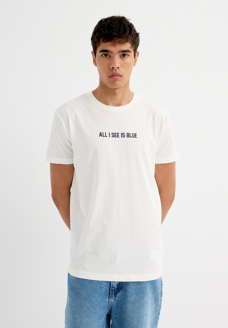Camiseta de algodón en color blanco roto con mangas cortas, escote redondo y el texto en marino "TODO LO QUE VEO ES AZUL" en el frente. Combinada con pantalones vaqueros azules.
