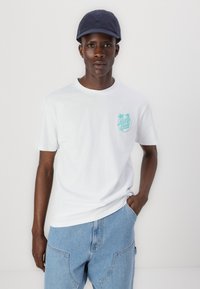 White cotton t-shirt with a turquoise graphic featuring palm trees on the left chest. Light blue denim pants and a navy cap.
