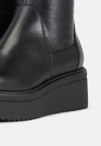 Botín negro de cuero hasta el tobillo con suela de plataforma gruesa, con detalles de costura y un acabado suave. Adecuado para varios estilos.